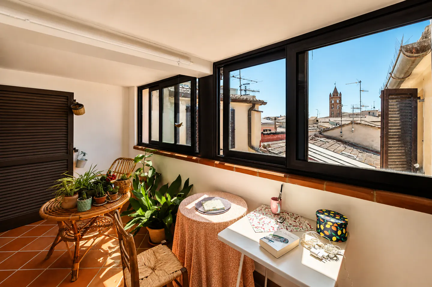 Bright sunroom with terracotta tile floor, wicker furniture, and a view of a European city through black-framed windows.
