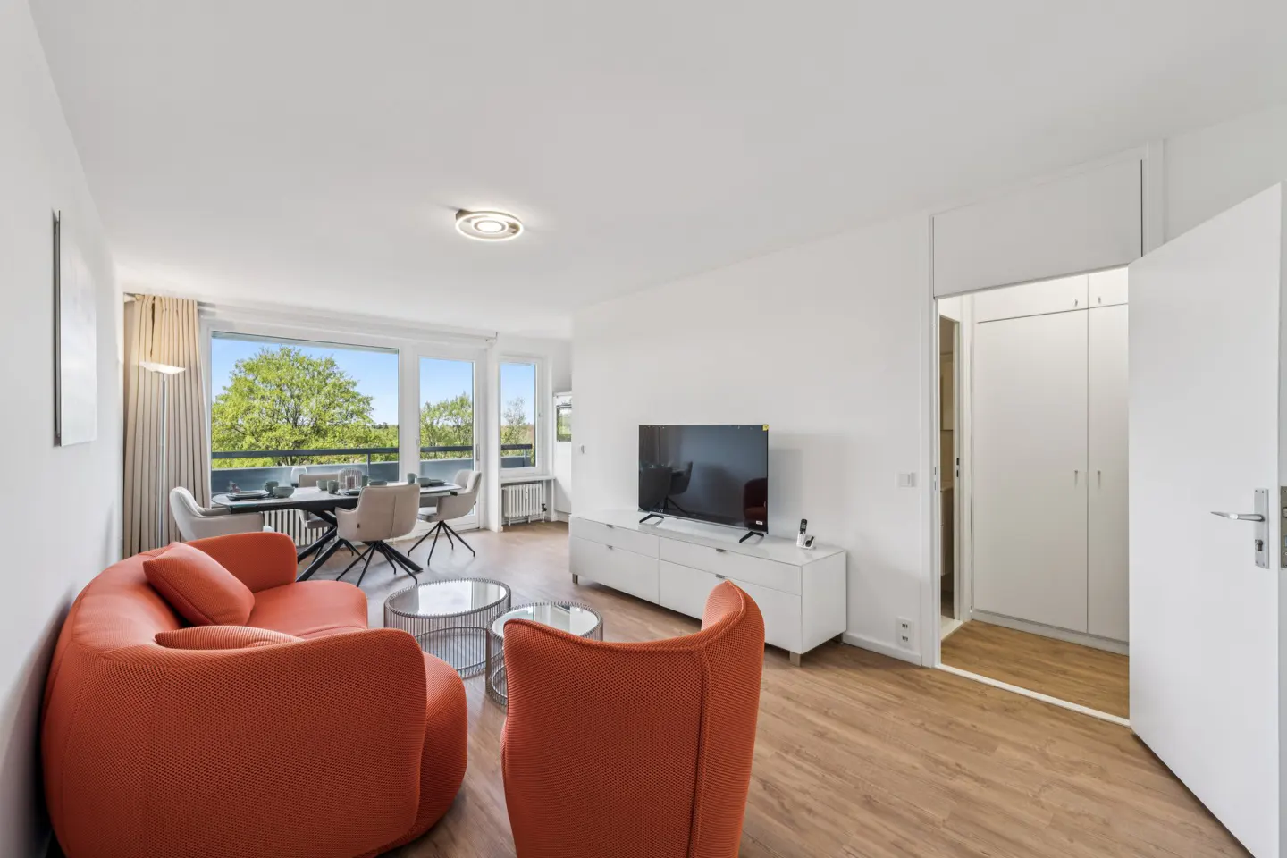 Bright living room with orange curved sofa, glass tables, and a dining area by a large window with a green view. A TV sits on a white cabinet.