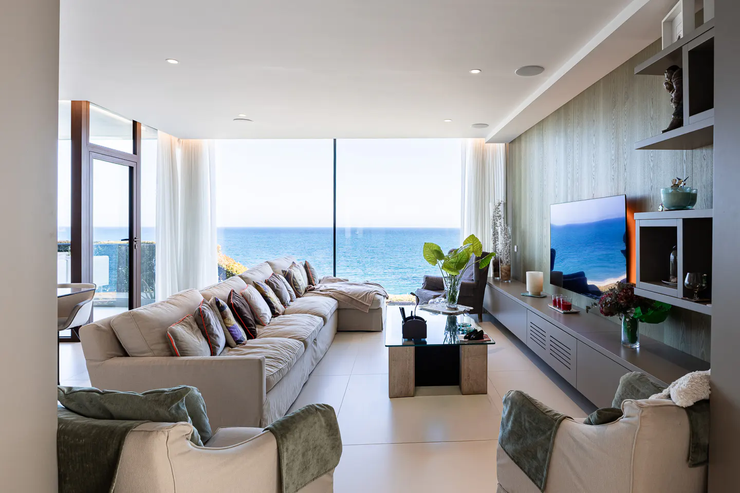 Bright living room with ocean view. Beige sofa with pillows, two armchairs, glass table, and TV on a gray cabinet.