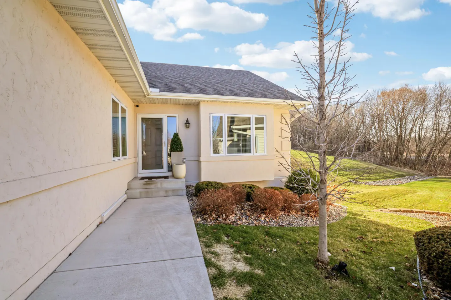 Beige house exterior with a gray roof, concrete walkway, and a tree in the front yard. Blue sky with clouds.