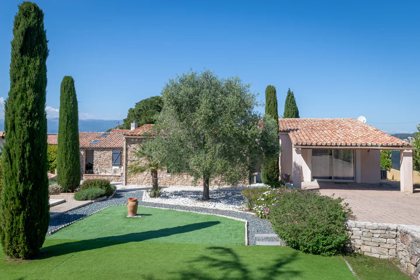 Exterior view of a stone house with a red tile roof, green lawn, trees, and blue sky.