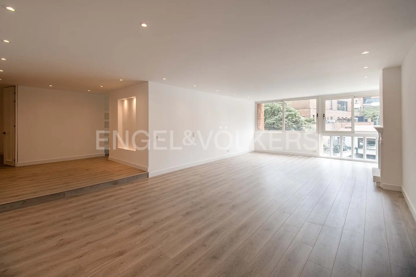 Bright, empty room with light wood floors, white walls, and recessed lighting. Large windows offer a city view.