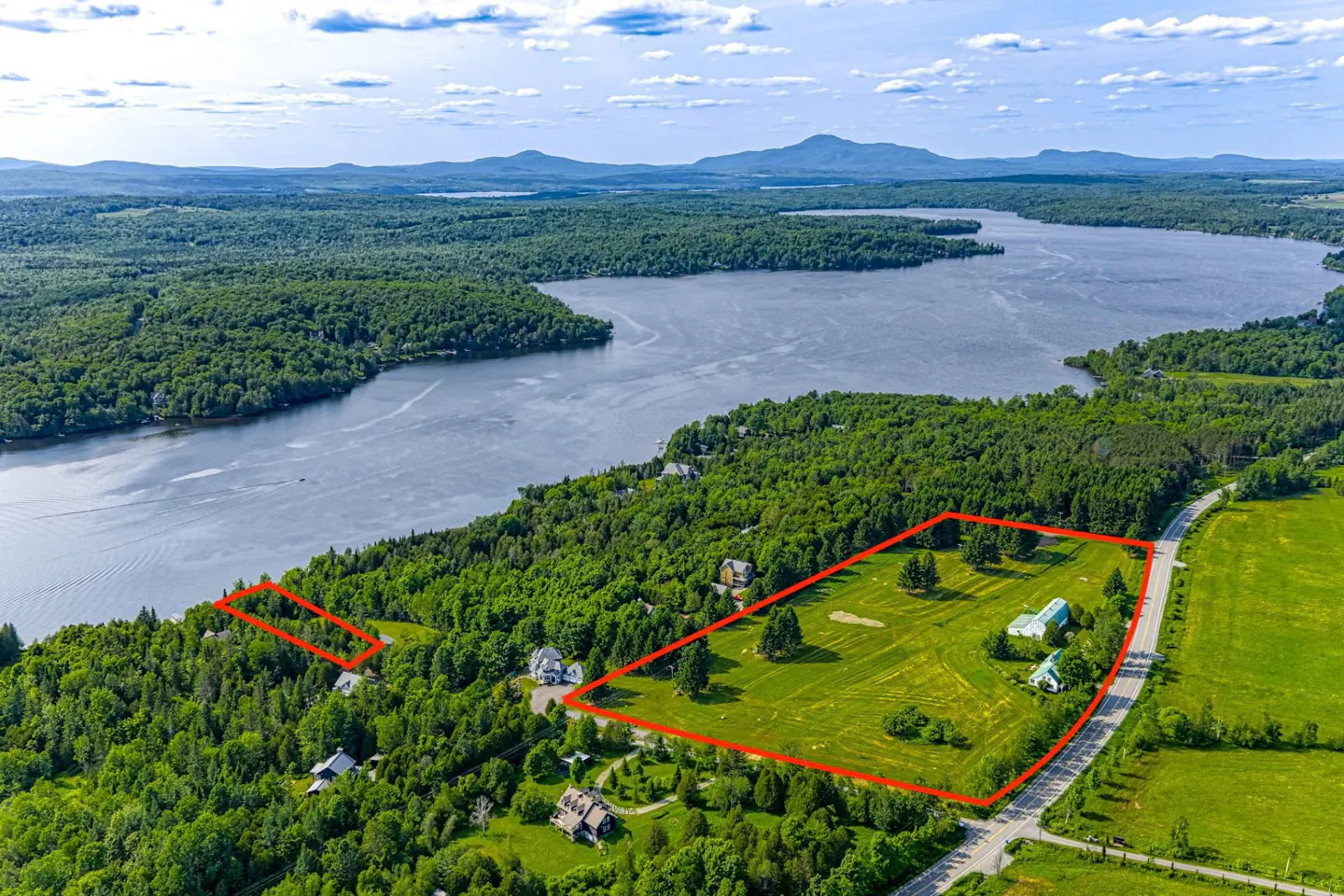 Aerial view of a large property outlined in red, featuring a lake, mountains, and lush greenery. Several houses are visible on the property.