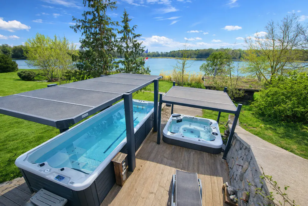 Outdoor swim spa and hot tub on a wooden deck with a lake view. Both are covered by a gray canopy.