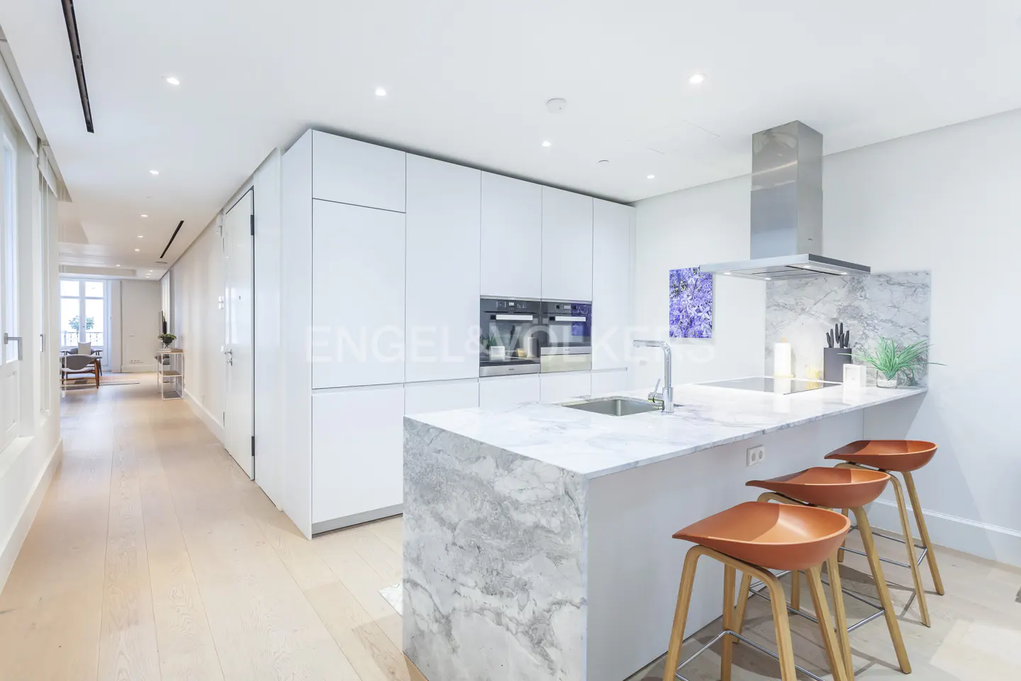 Bright, modern kitchen with white cabinets, marble island with three brown stools, and stainless steel appliances. Hallway leads to a sunlit room.