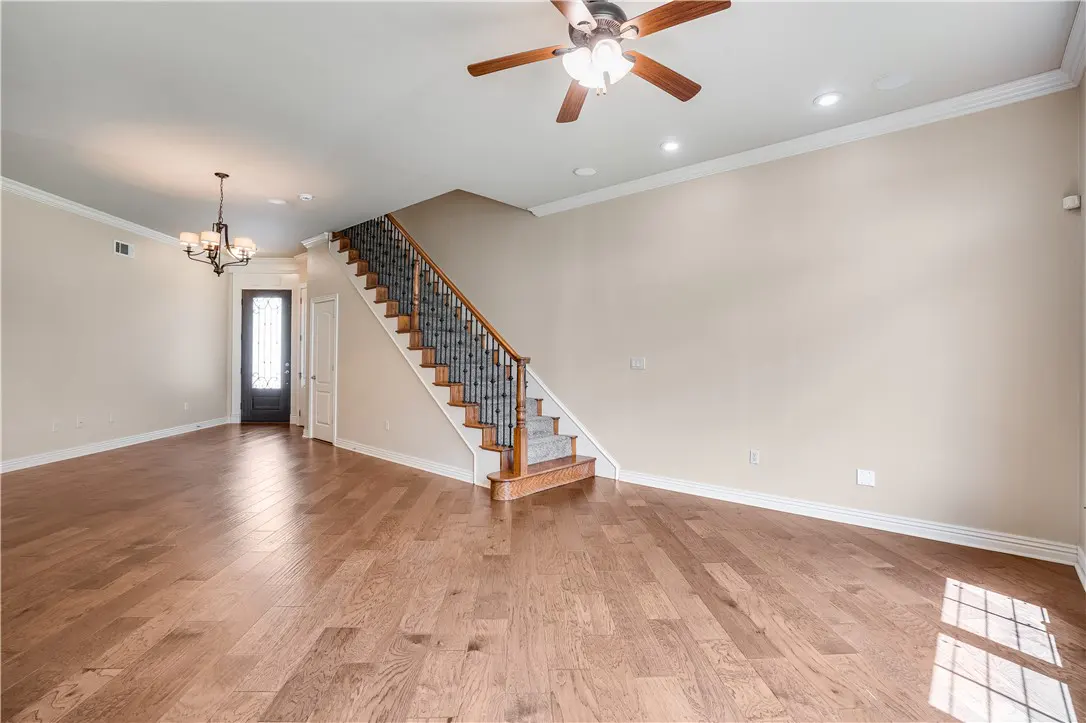 A bright, empty living room with hardwood floors, beige walls, and a staircase with a decorative iron railing. A ceiling fan and chandelier provide light.