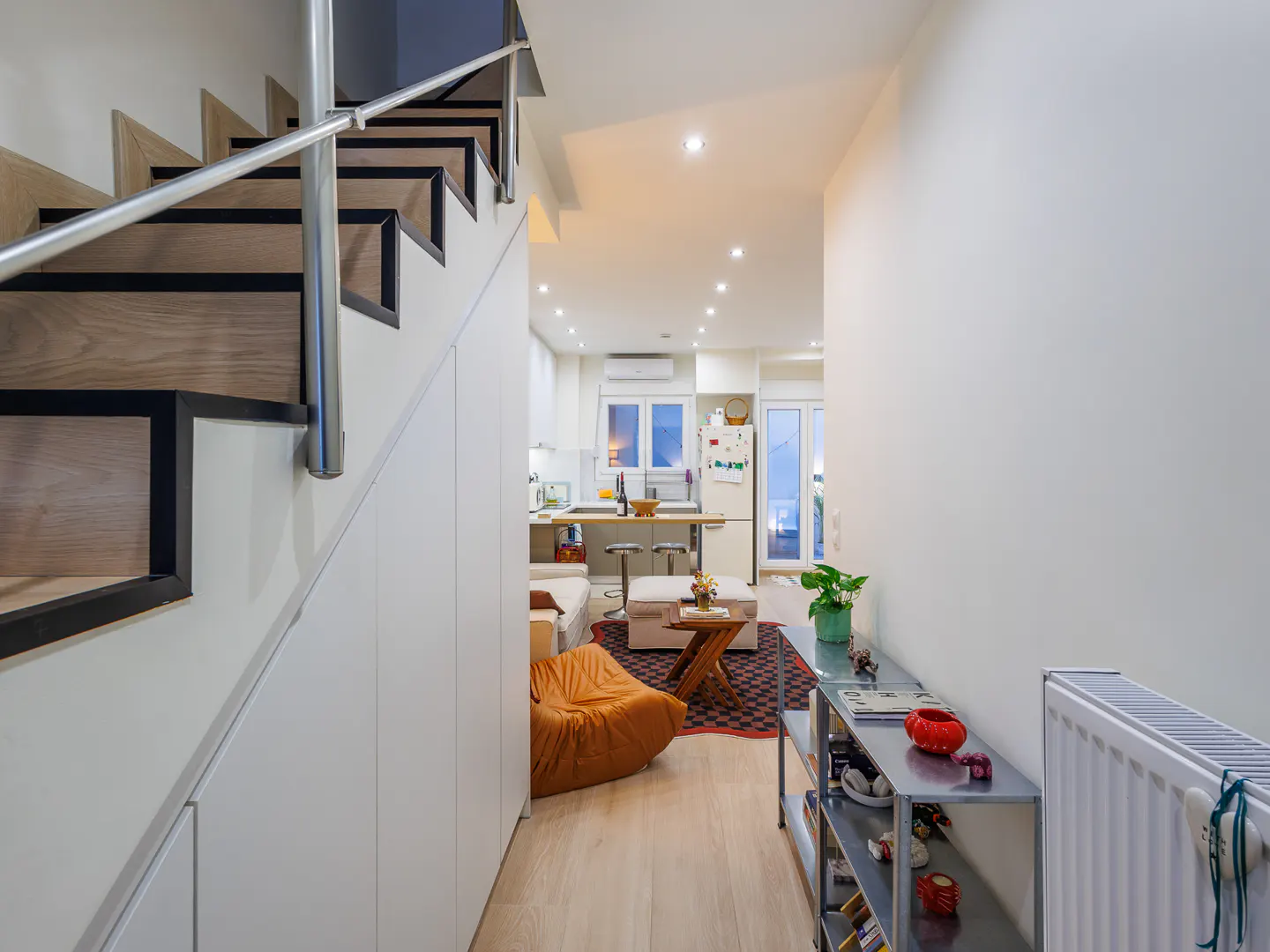 Interior view of a modern apartment with stairs, white walls, and light wood floors. A living area with a kitchen is visible in the background.