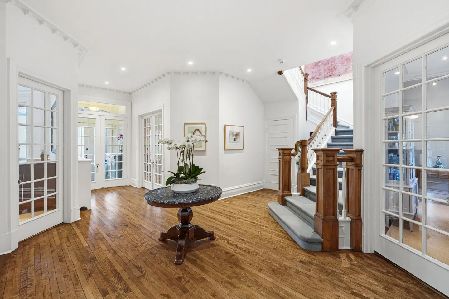 Bright foyer with hardwood floors, white walls, and a staircase. A round table with orchids sits in the center. French doors lead to other rooms.