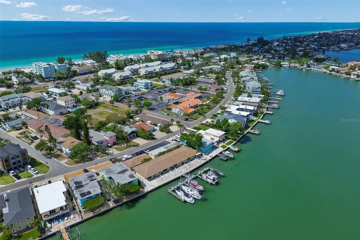 Aerial view of a coastal neighborhood with houses, docks, boats, and a blue ocean under a partly cloudy sky.
