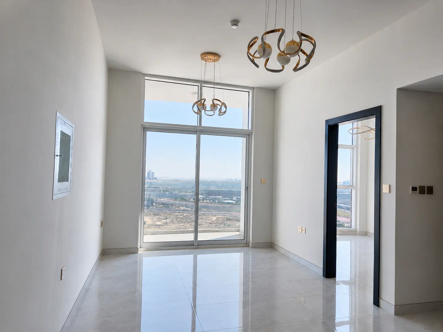 Bright, empty room with white walls, shiny tile floor, and city view through a large window. Modern light fixtures hang from the ceiling.