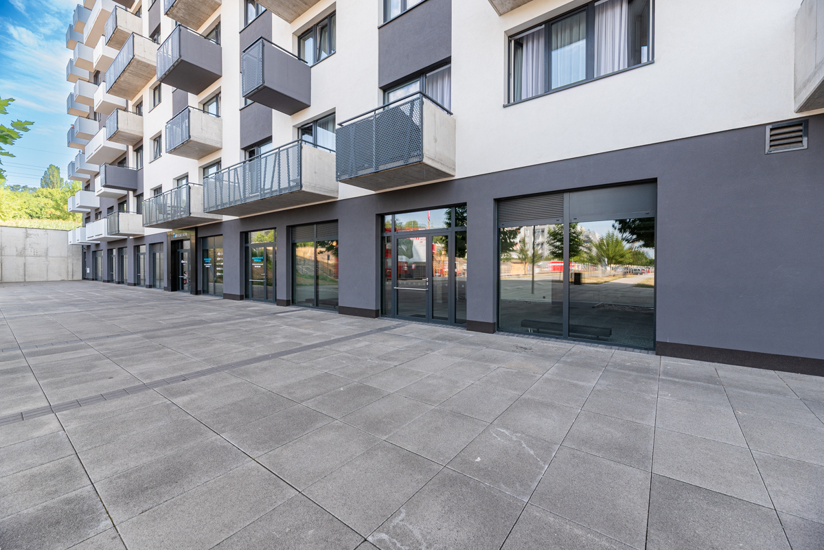 Modern apartment building with white and grey facade, balconies, and ground-floor commercial storefronts on a paved plaza. [AI-generated text]
