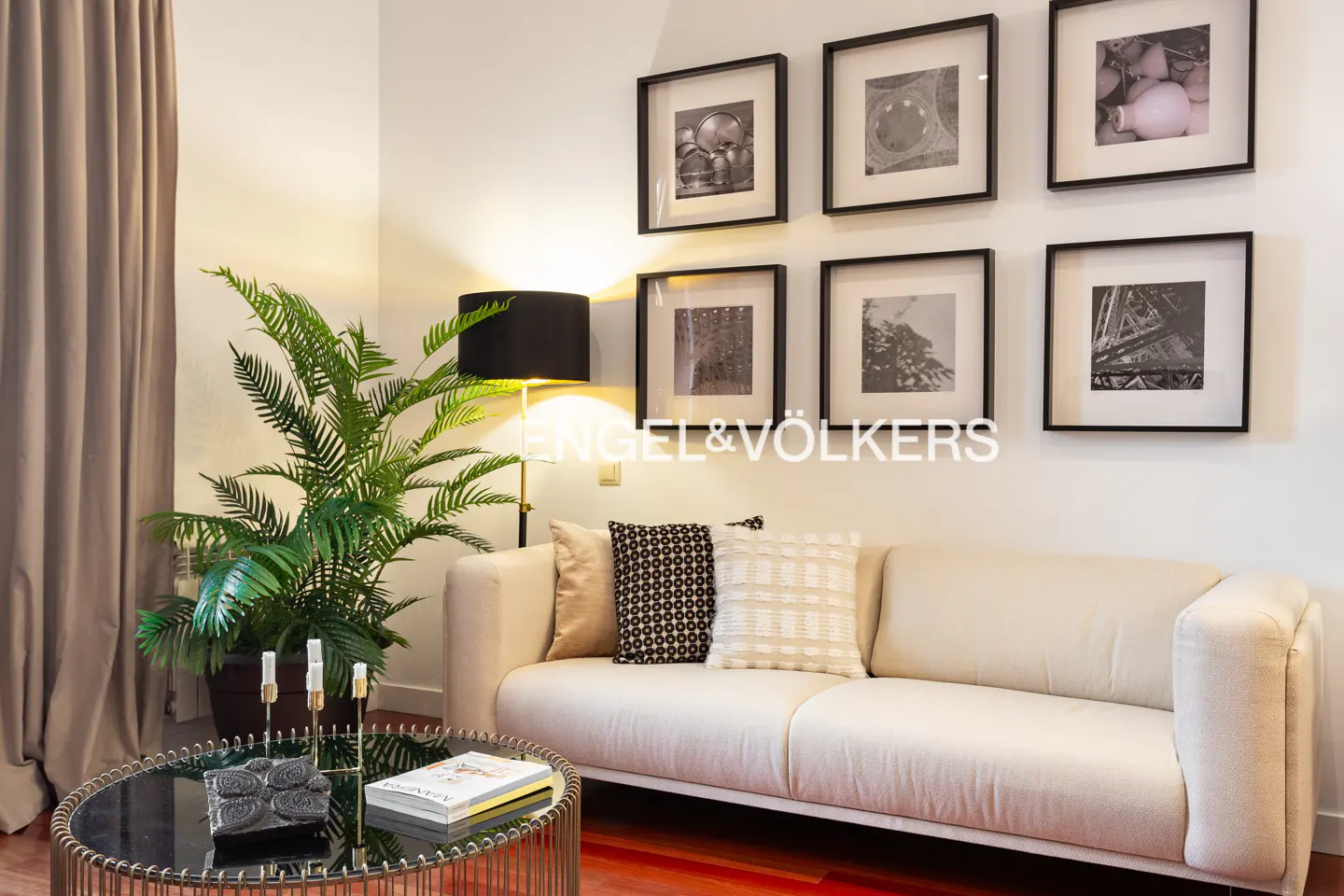 A beige sofa with patterned pillows sits next to a potted plant, a floor lamp, and a gallery wall of framed photos.