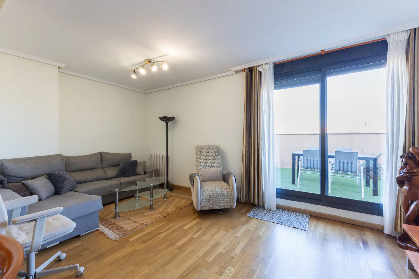 Living room with a gray sectional sofa, patterned armchair, and glass table. A sliding glass door leads to a balcony with outdoor furniture.
