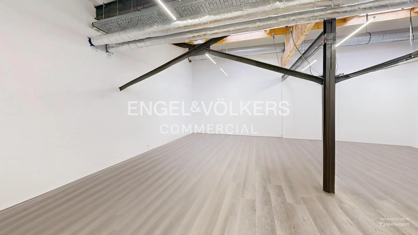 Empty commercial space with white walls, gray wood-look floors, and exposed ceiling with metal support beam. Engel & Völkers logo on wall.