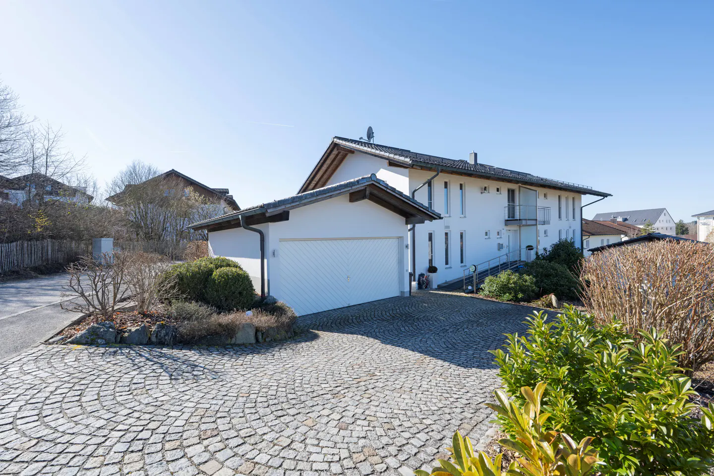 Exterior of a two-story white house with a garage and a circular cobblestone driveway. Shrubs and trees surround the property.