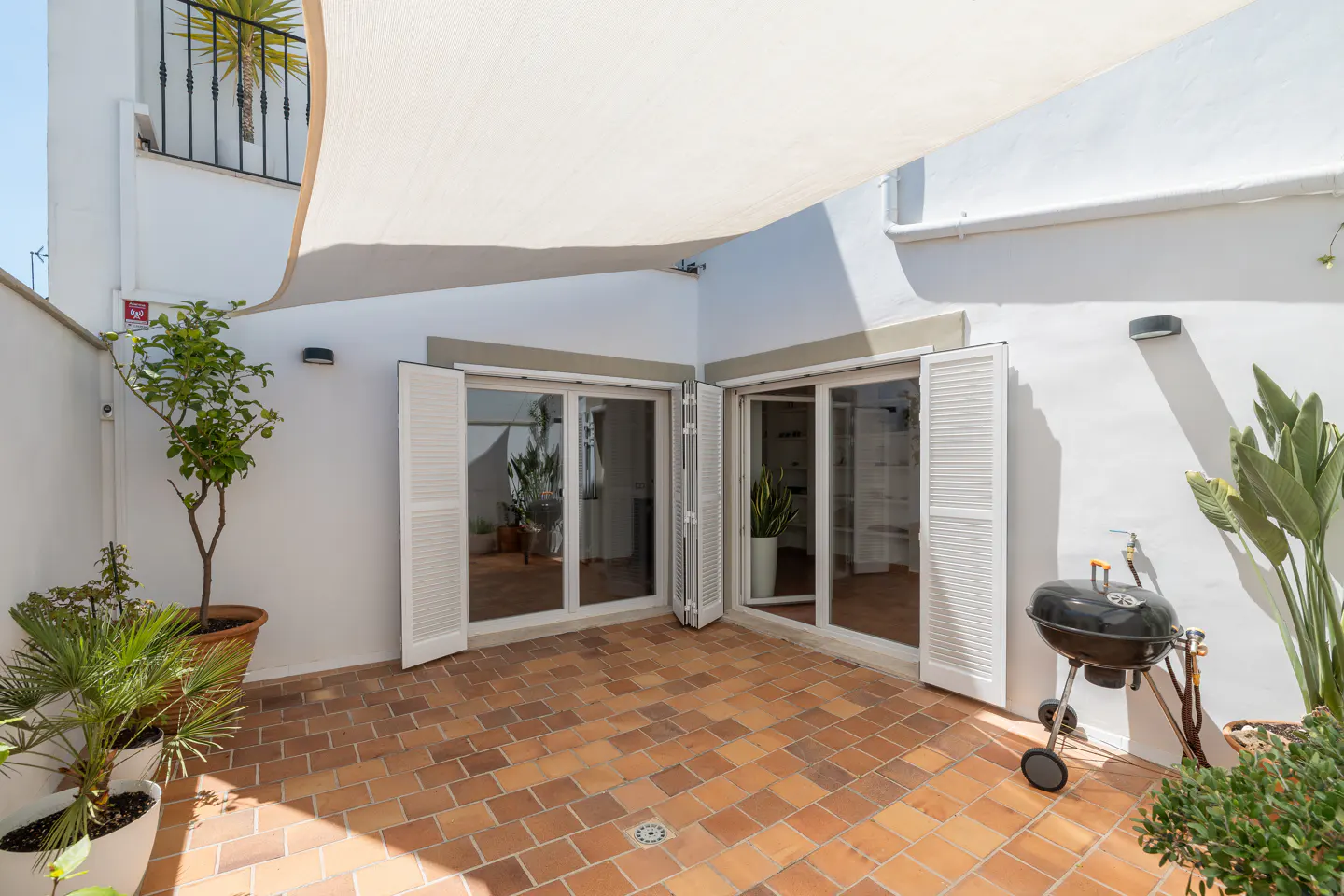 Outdoor patio with terracotta tiles, white walls, and open shuttered doors. Plants in pots, a grill, and a white awning provide shade.