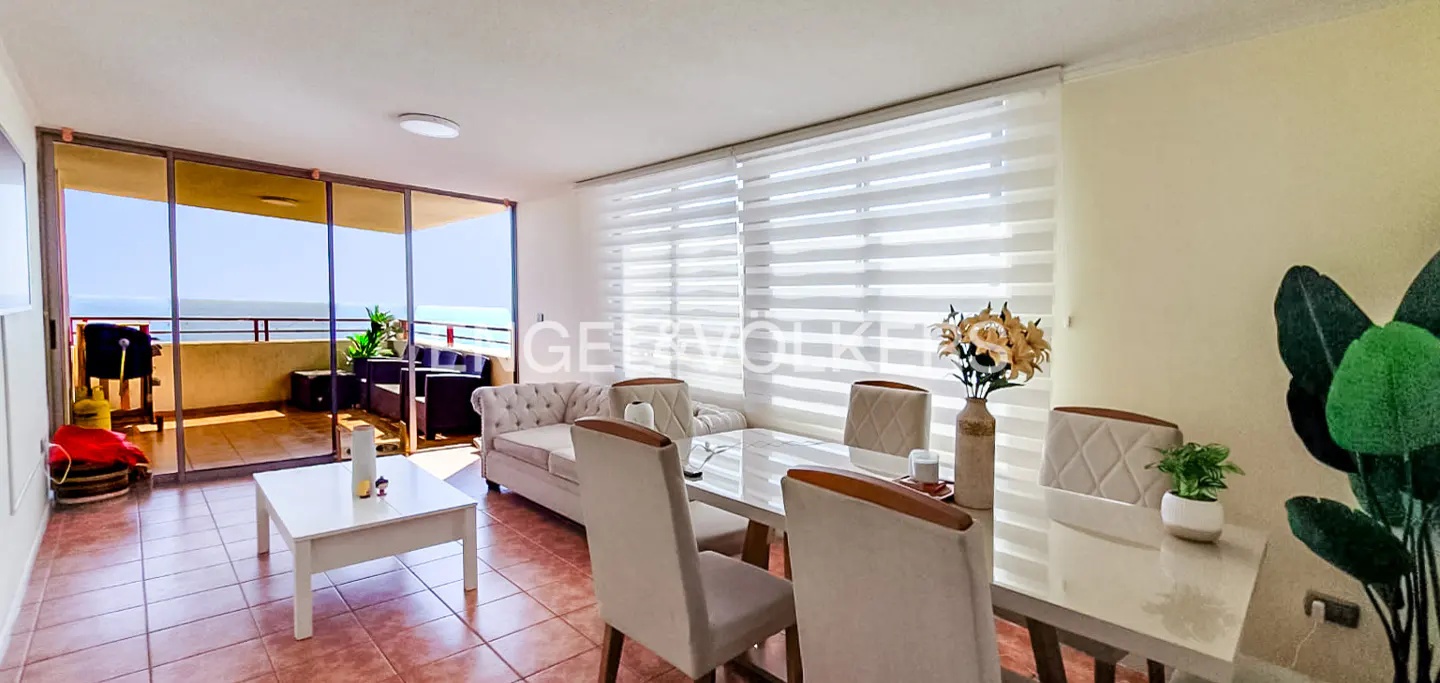 Bright living room with a balcony overlooking the ocean. A white table and chairs are in the foreground.