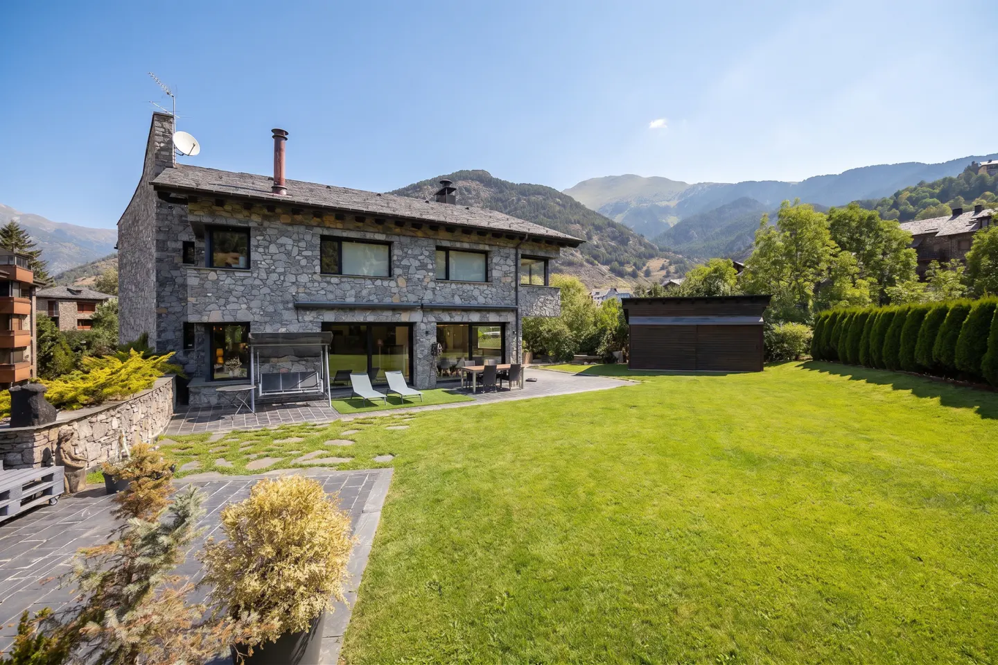 Two-story stone house with a green lawn, patio furniture, and mountain views under a clear blue sky.