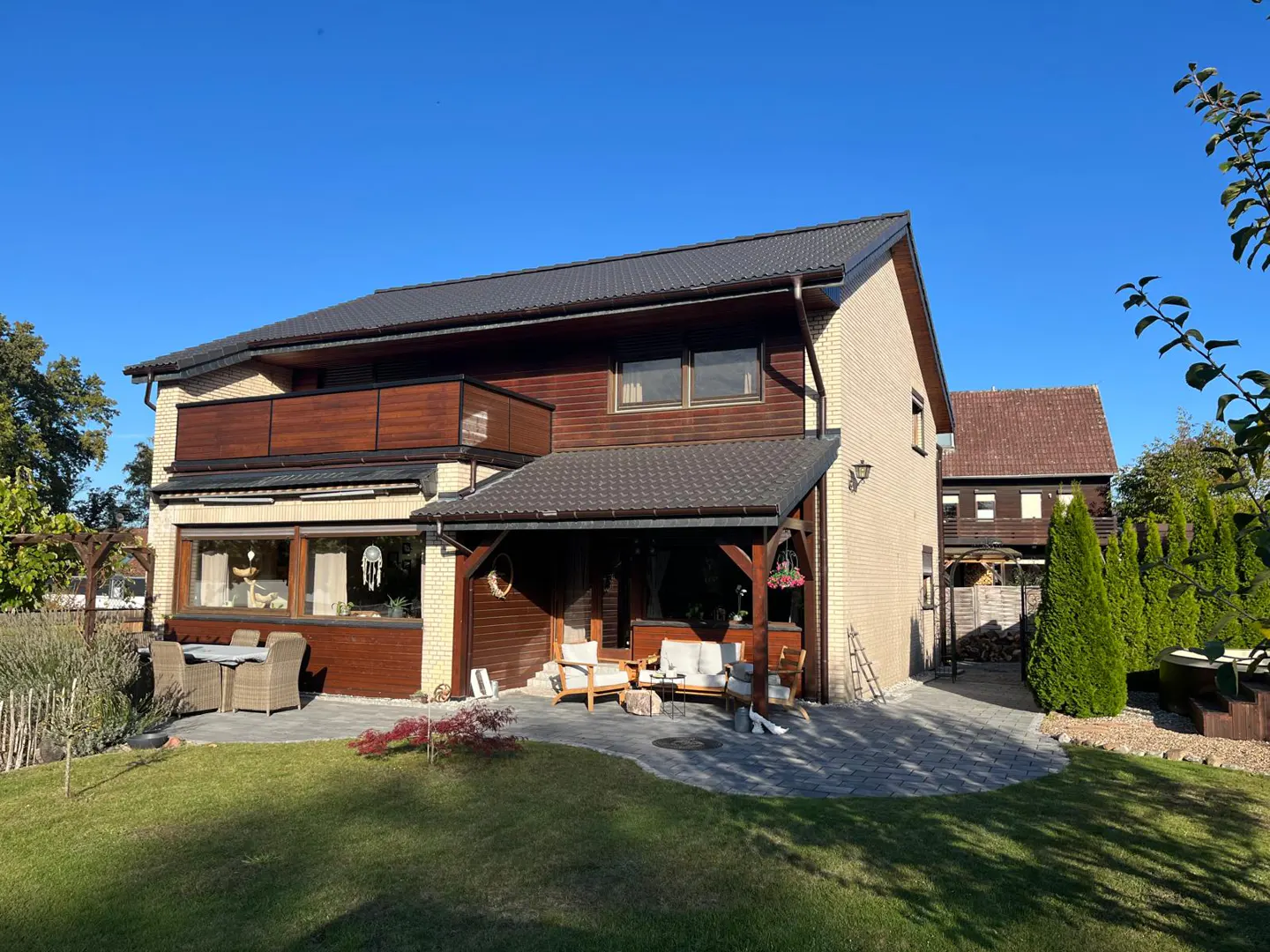 Two-story house with a brown roof, brick and wood siding, and a patio with outdoor furniture. Green lawn and blue sky.