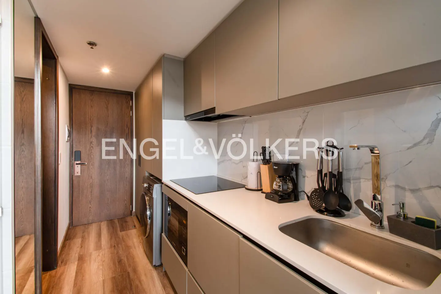 Modern kitchen with gray cabinets, white marble backsplash, stainless steel sink, and wood-look flooring. Doorway visible on the left.