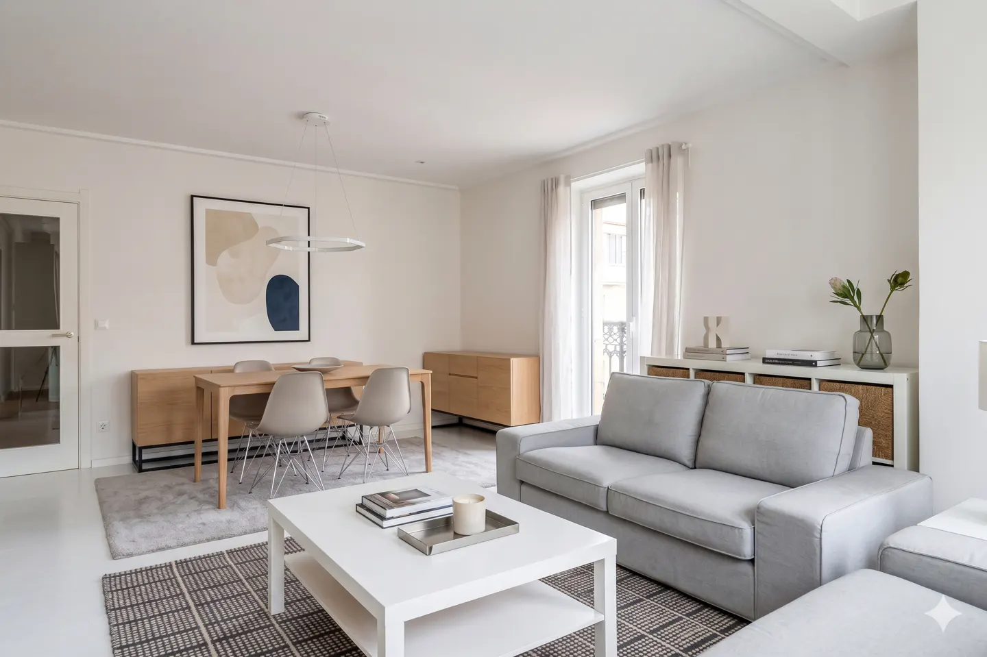 Bright, modern living room with a gray sofa, white coffee table, and wooden dining set. Abstract art hangs above the dining table.