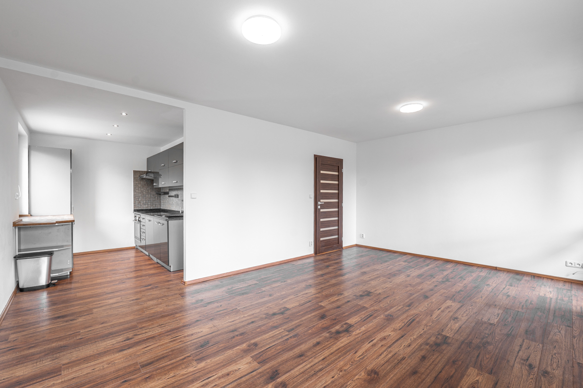 Empty room with dark wood floors, white walls, a brown door, and a view into a modern kitchen with grey cabinets.