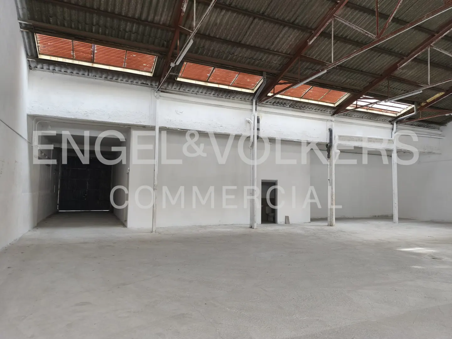 Empty commercial space with white walls, concrete floor, and metal roof with skylights. Engel & Volkers Commercial logo is superimposed.
