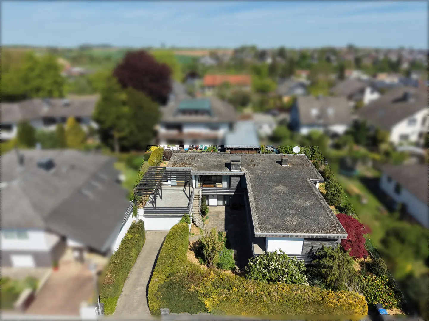 Aerial view of a modern, two-story house with a flat roof, a pergola-covered patio, and a long driveway lined with green hedges.