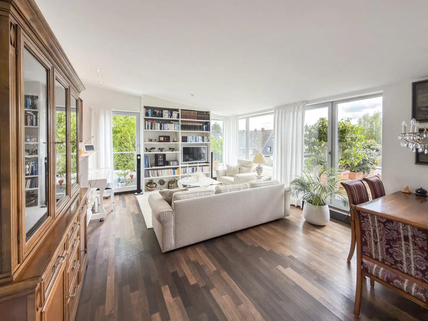 Bright living room with dark wood floors, a white sofa, a large bookshelf, and floor-to-ceiling windows with garden views.
