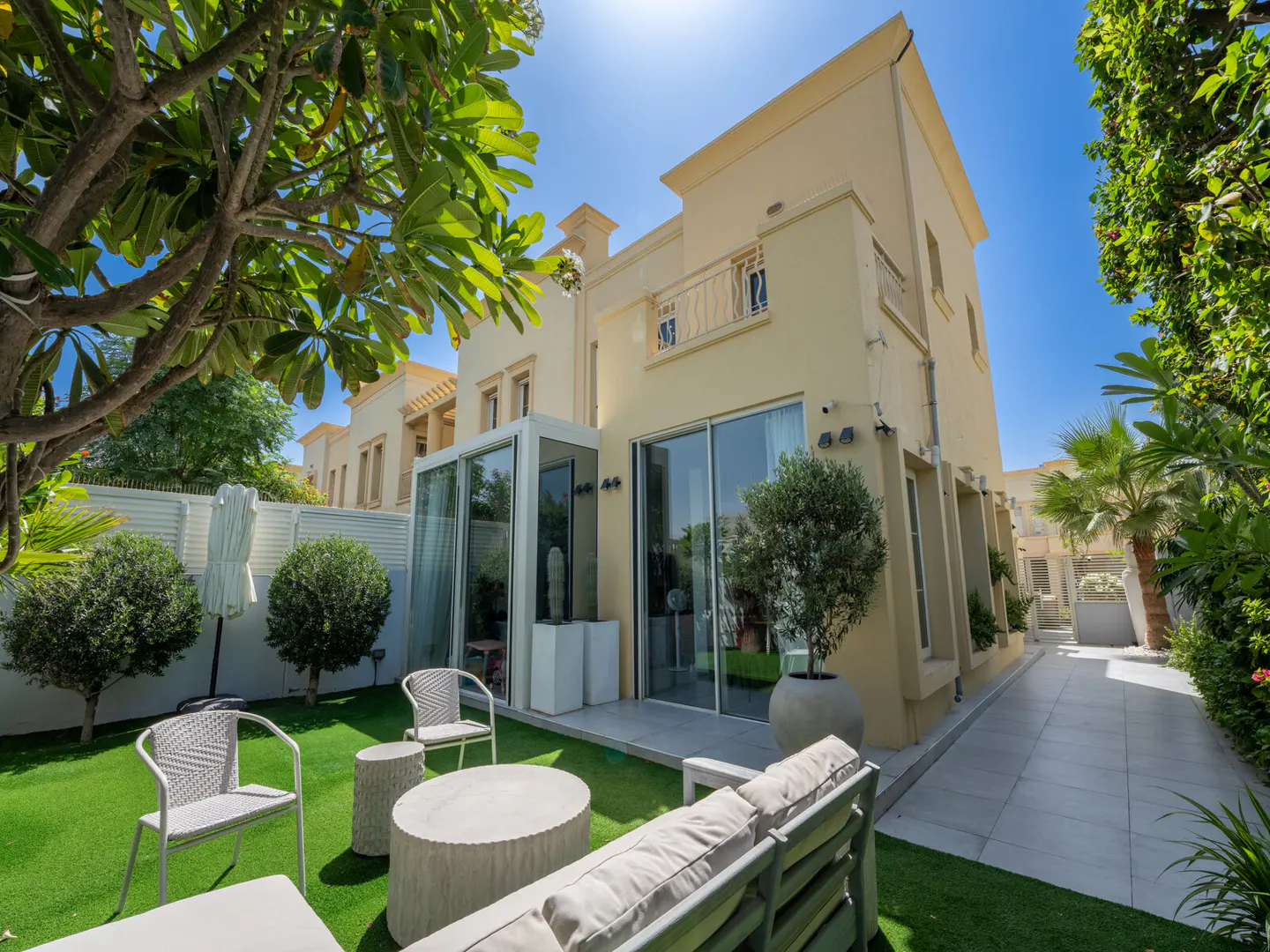 Backyard view of a two-story beige house with a patio set on artificial grass, and a tiled walkway.