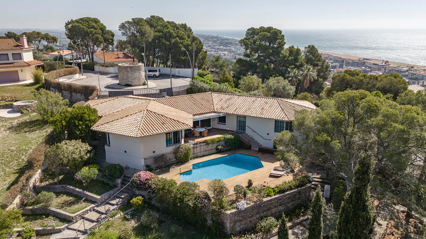 Aerial view of a white house with a terracotta tile roof and a blue swimming pool, surrounded by green trees and a view of the ocean.