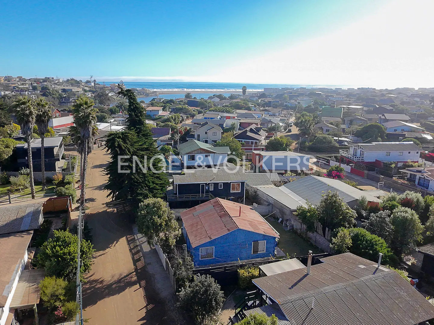 Aerial view of a coastal town with blue houses, palm trees, and the ocean in the background under a clear blue sky.