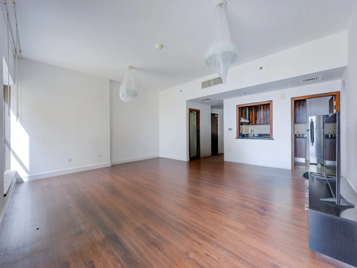 Bright, empty apartment with wood floors, white walls, and a kitchen visible through an opening. Chandeliers are covered in plastic.