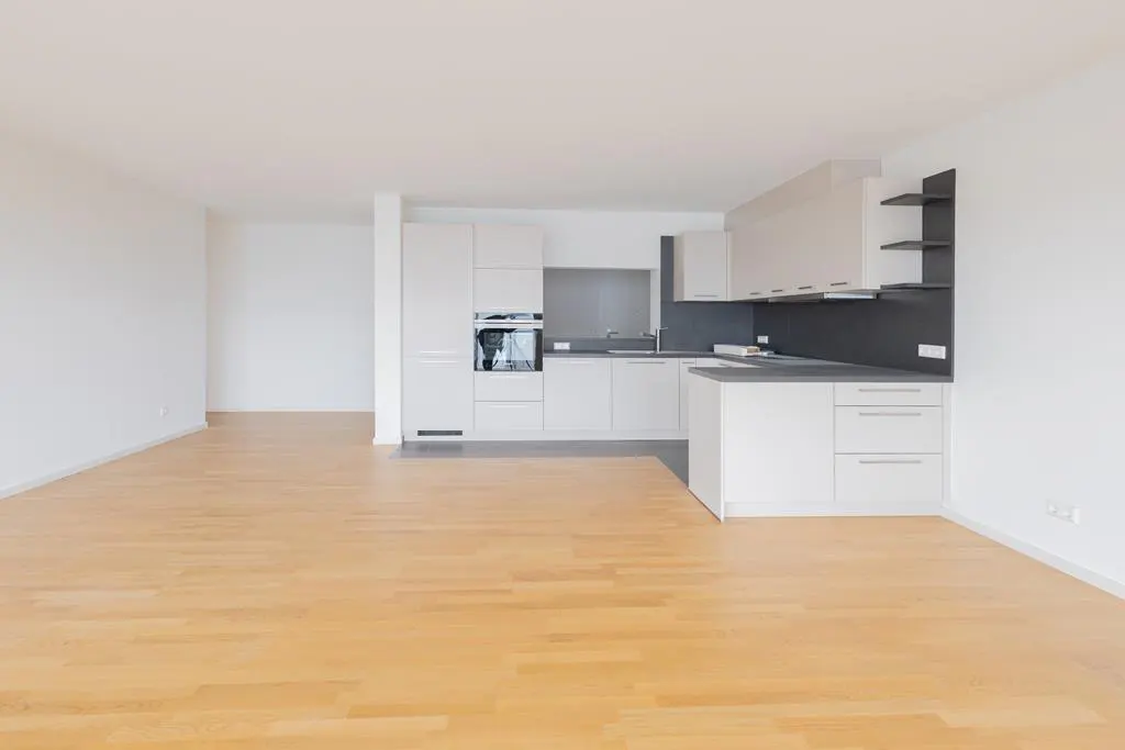 Bright, empty apartment with light wood floors and white walls. A modern kitchen with white cabinets and dark backsplash is in the background.