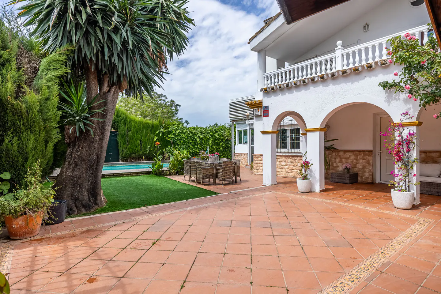 Outdoor patio with terracotta tiles, a dining table, and a pool surrounded by lush greenery and a white two-story house.