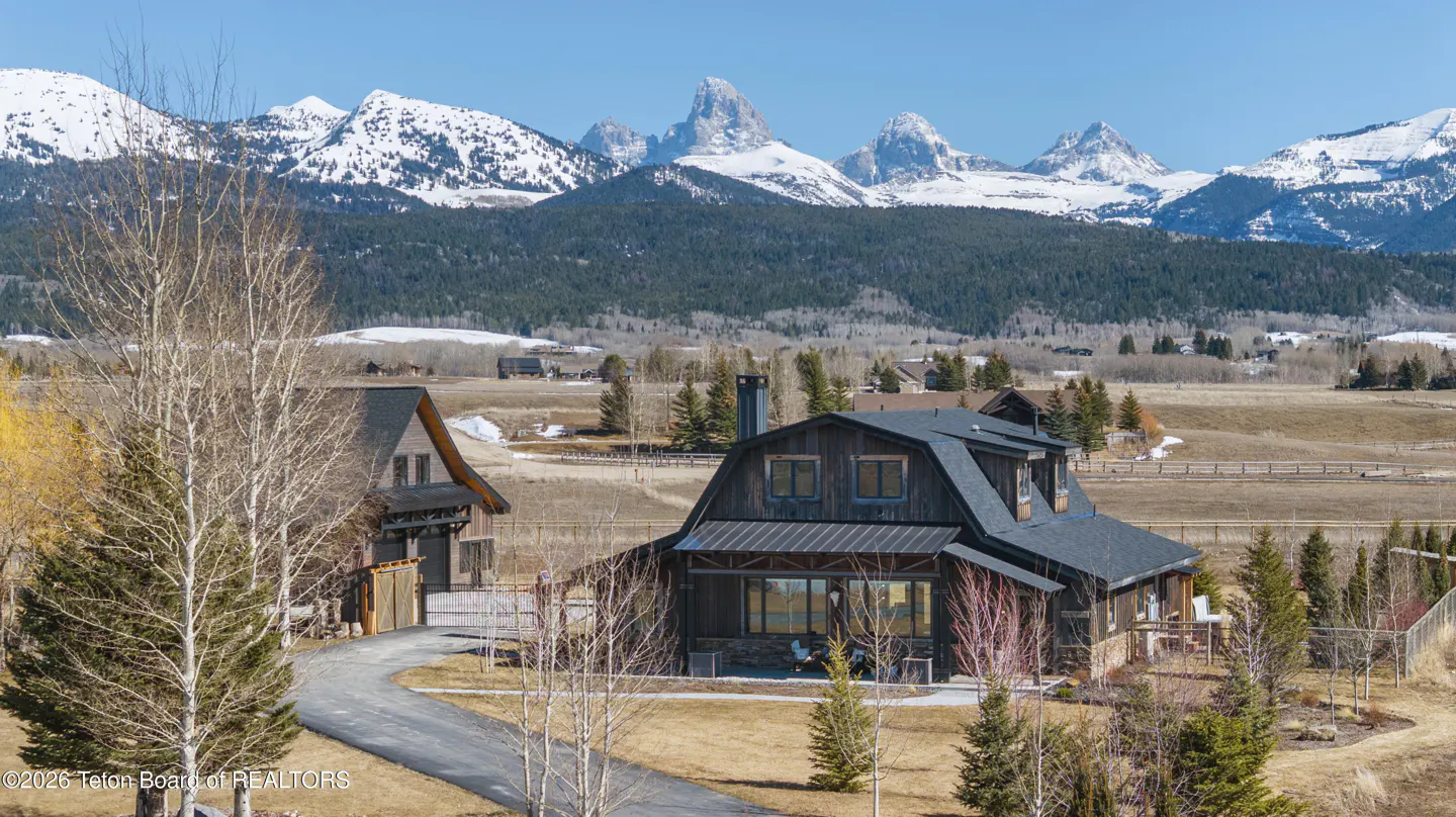A dark brown house with a black roof sits before snow-capped mountains under a blue sky.