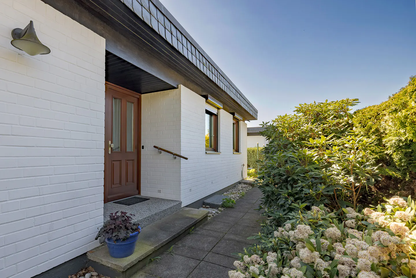 Exterior view of a white brick house with a brown front door and a walkway lined with greenery.