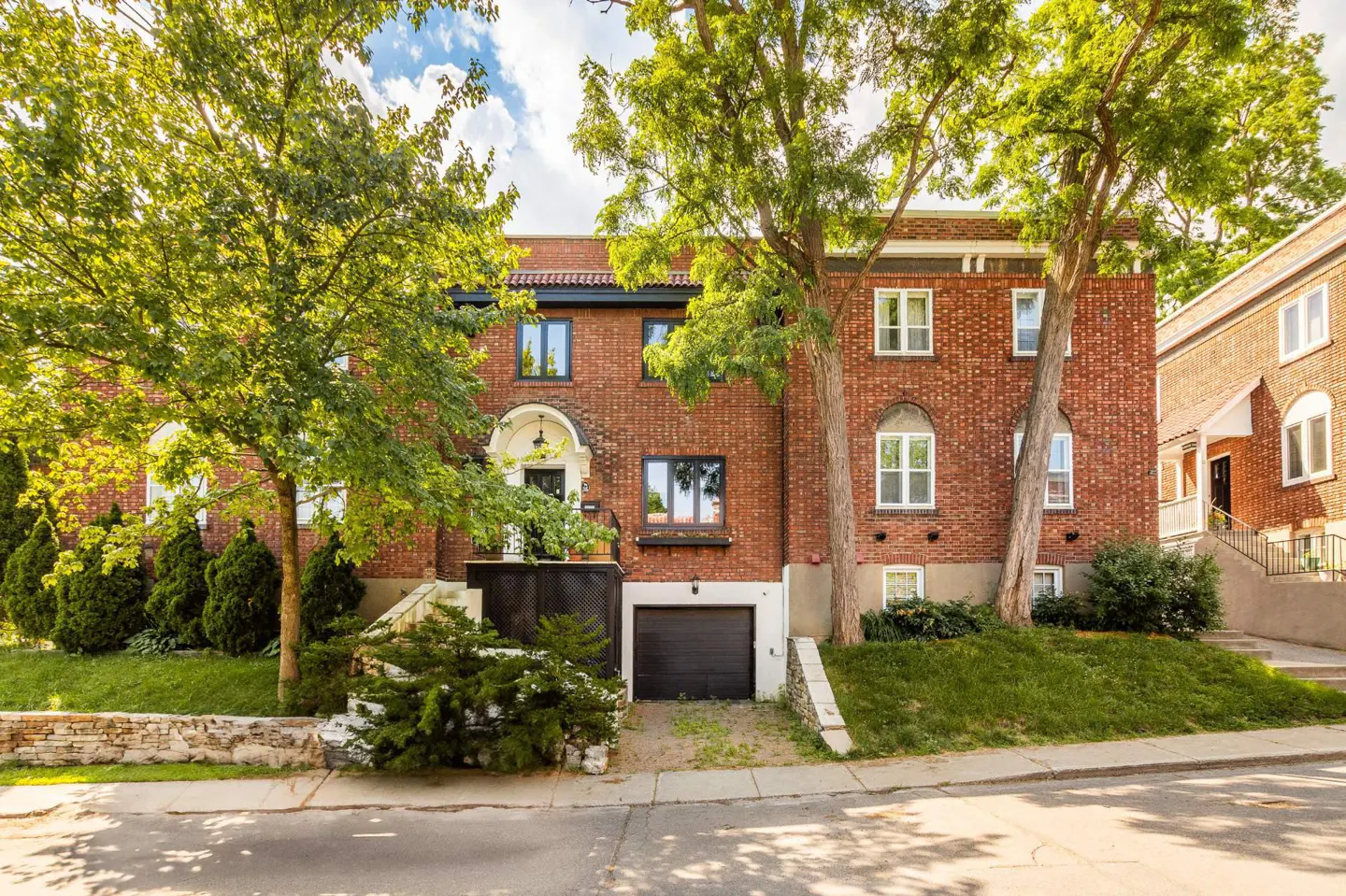 A three-story red brick house with a black garage door and green trees in the front yard.