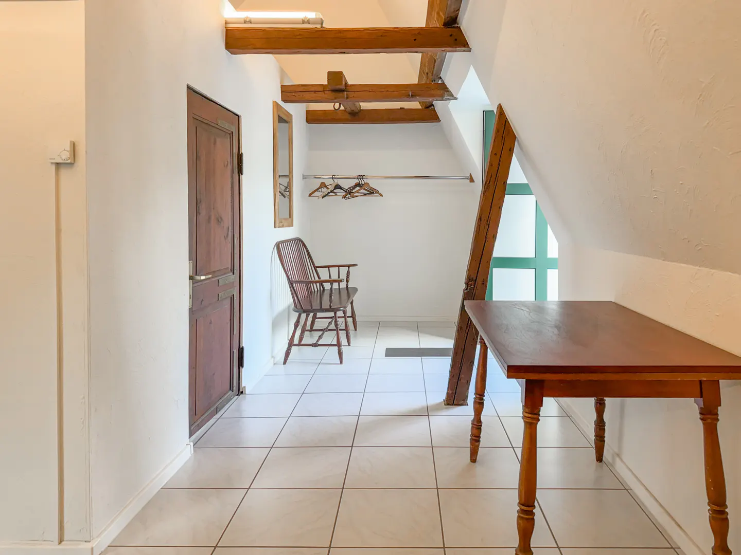 Attic room with white walls, tile floor, and exposed wooden beams. A brown wooden door, chair, and table are visible.