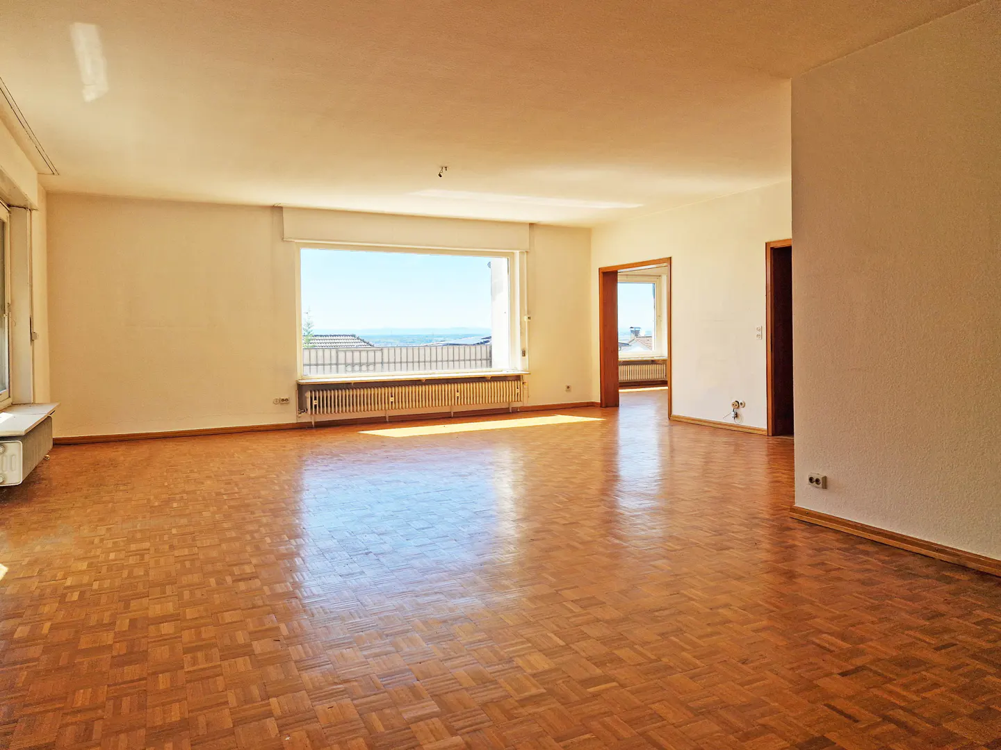 Spacious, empty living room with parquet floors, white walls, and a large window with a view of the sea.
