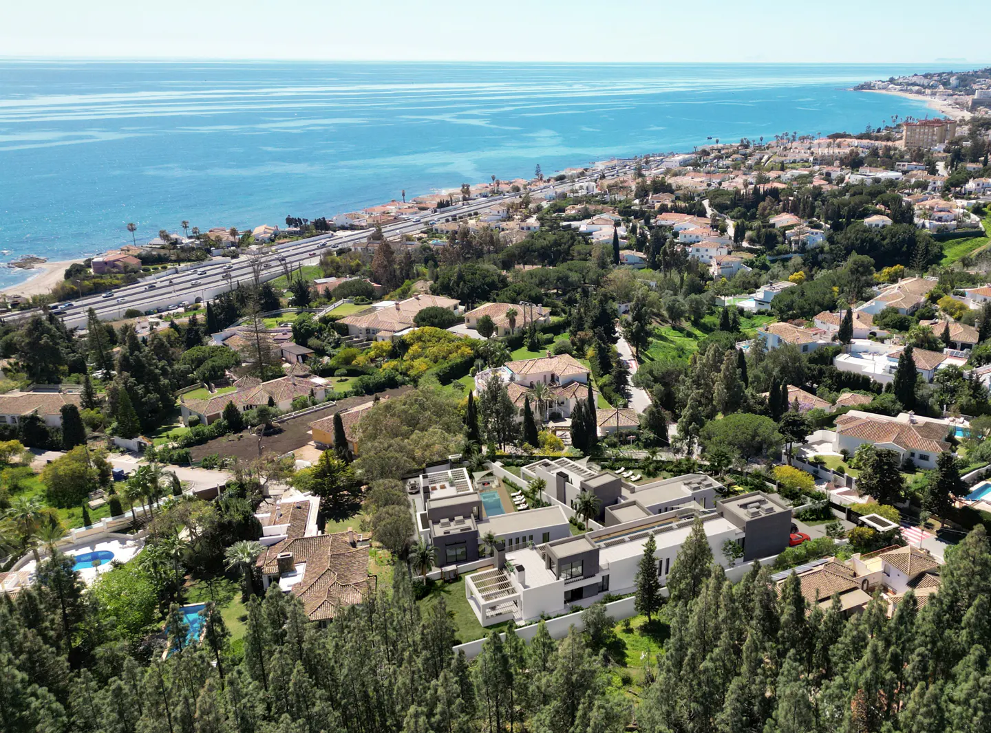 Aerial view of modern white villas with pools nestled among trees, with the blue ocean and coastline in the background.