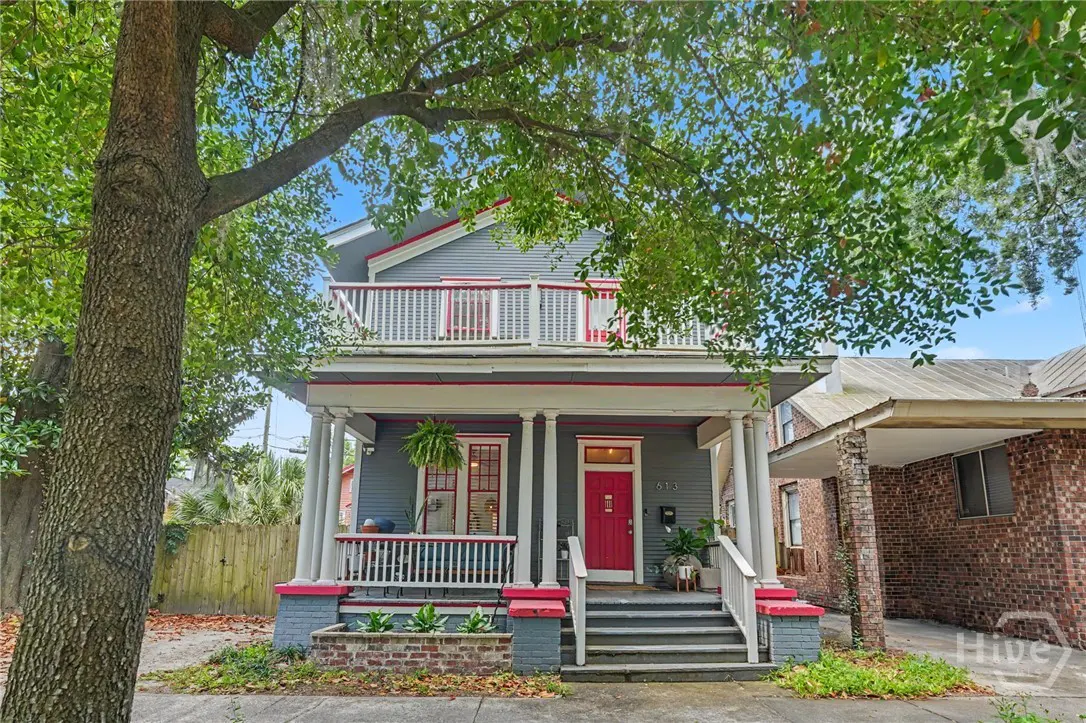 Two-story gray house with a red door and trim, white columns, and a second-story balcony. A large tree is in the front yard.