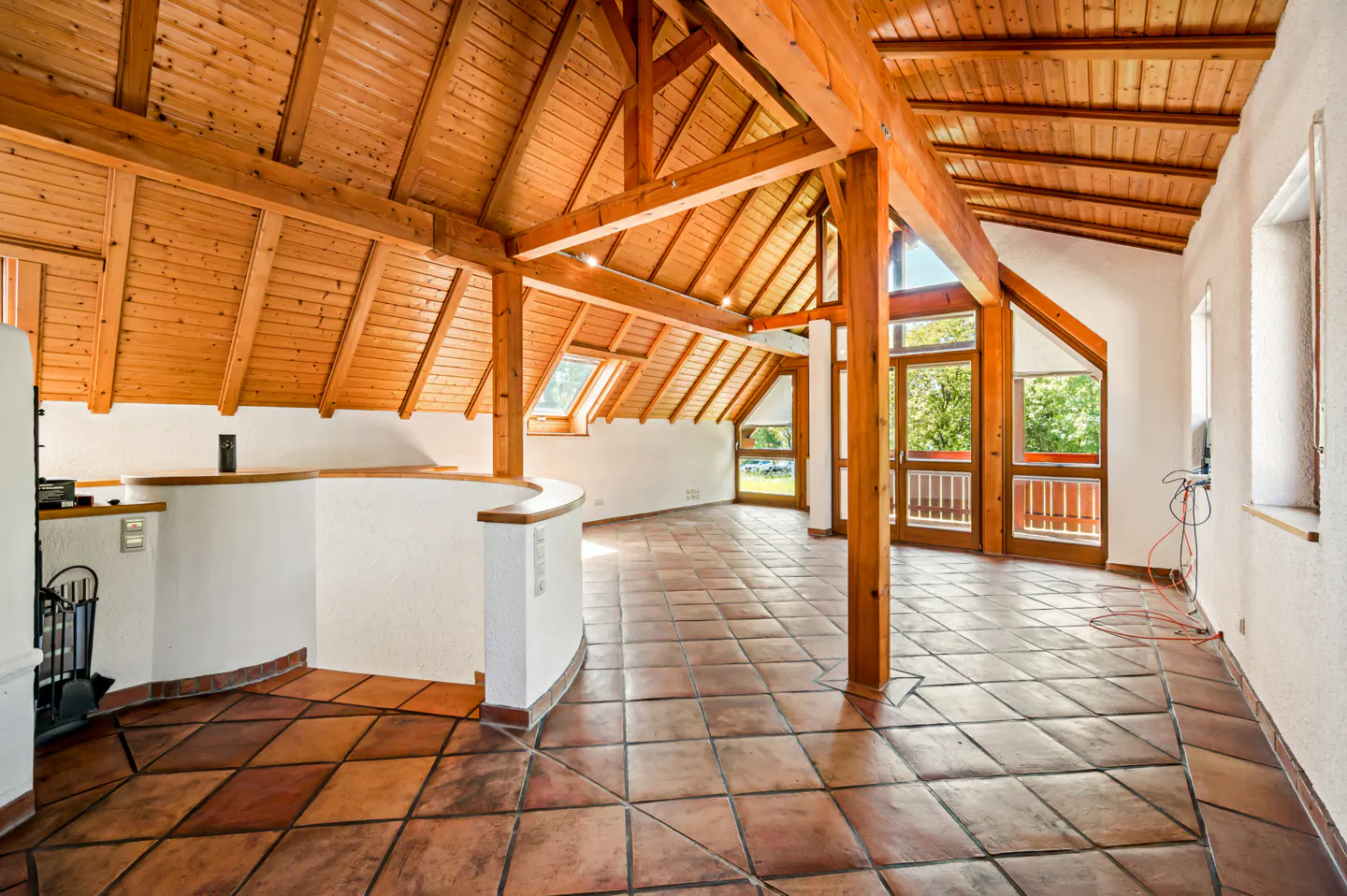 Attic room with exposed wood beams, terracotta tile floor, and white walls. Large windows and doors let in natural light.