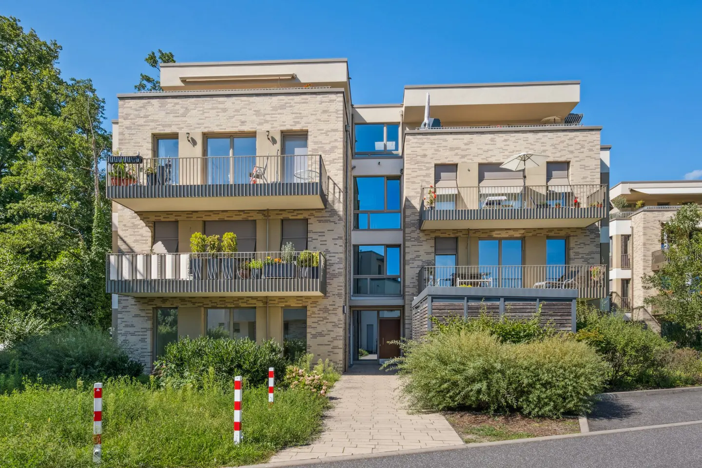 Modern apartment building with balconies, tan brick facade, and blue windows under a clear sky. Lush greenery surrounds the building.