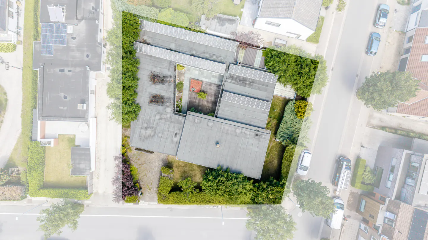 Aerial view of a flat-roofed house with solar panels, a central courtyard, and surrounding greenery in a residential area.