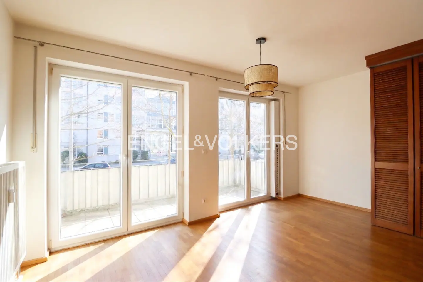 Bright, empty room with hardwood floors, white walls, and large windows. A straw chandelier hangs from the ceiling. A wooden closet is on the right.