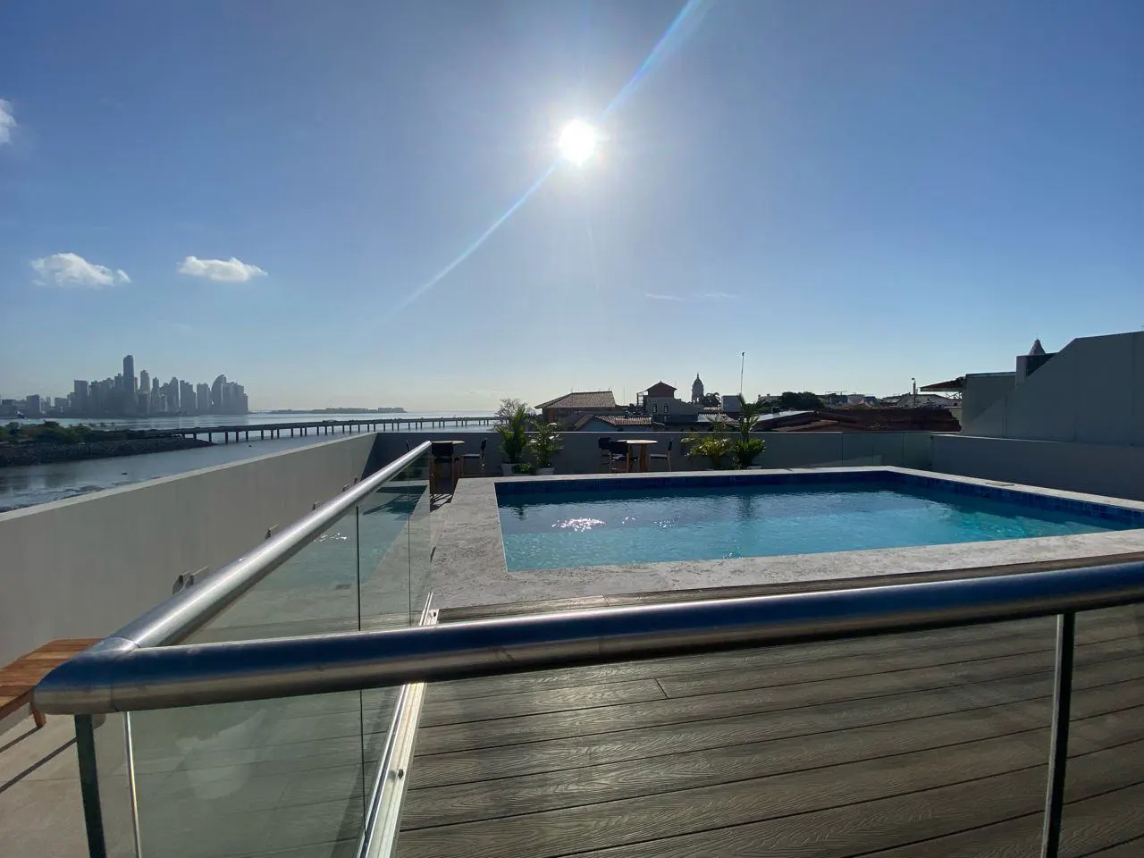 Rooftop pool with wood deck and glass railing. City skyline and bridge in the distance under a bright blue sky.