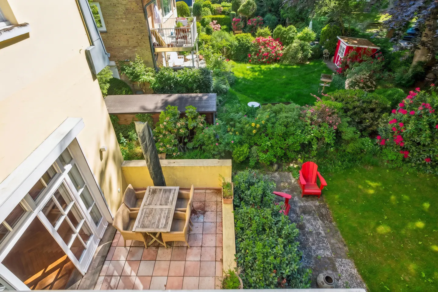 Aerial view of a patio with a table and chairs, leading to a lush green garden with red chairs and a shed.
