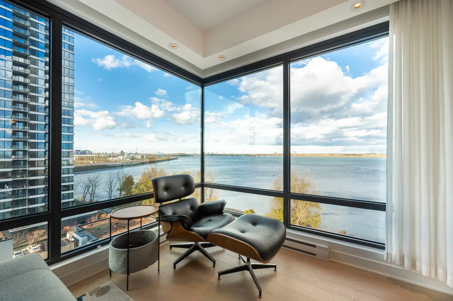 Corner view of a modern apartment with a black leather chair and ottoman, large windows overlooking a river and city skyline.