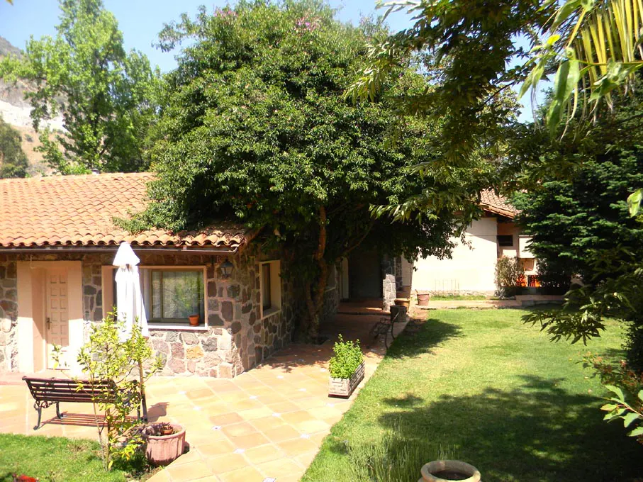 Exterior view of a stone house with a red tile roof, surrounded by green trees and a lawn. A bench sits on a tiled patio.